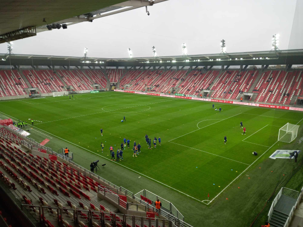 Ein Fußballfeld in einem Stadion mit Menschen, Stühlen, Geländern, Treppen und Lampen, unter einem offenen Himmel.