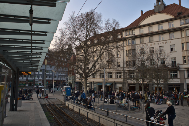 Eine belebte Straßenkreuzung mit einem Bahnhof auf der linken Seite, umgeben von Gebäuden, Bäumen und Laternen, mit Menschen, die gehen, auf Bänken sitzen und in der Nähe des Bahnhofs stehen.