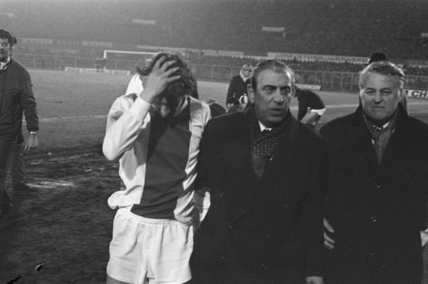 Eine Gruppe von Männern in Bedrängnis steht zusammen auf einem Fußballfeld, mit einem Stadion im Hintergrund, auf einem Schwarz-Weiß-Bild.