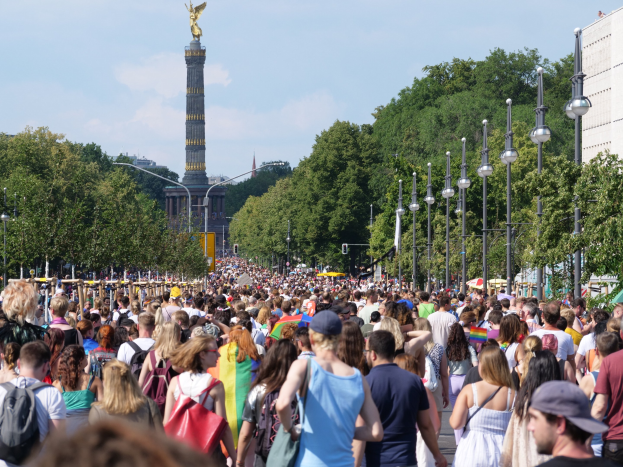 Eine große Menschenmenge geht eine Straße in Berlin, Deutschland, entlang und trägt Taschen mit Laternen an der Straße, Bäumen und einem Turm mit einer Statue im Hintergrund, umgeben von Gebäuden unter einem bewölkten Himmel.