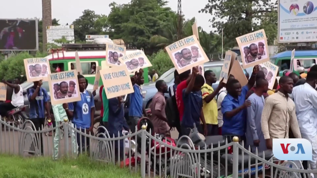 Eine Gruppe von Menschen h├Ąlt Schilder, wahrscheinlich protestierend, vor einem Zaun mit Fahrzeugen, B├Ąumen, Masten, Absperrungen und einem Bildschirm im Hintergrund, unter einem sichtbaren Himmel mit Gras am unteren Bildrand.