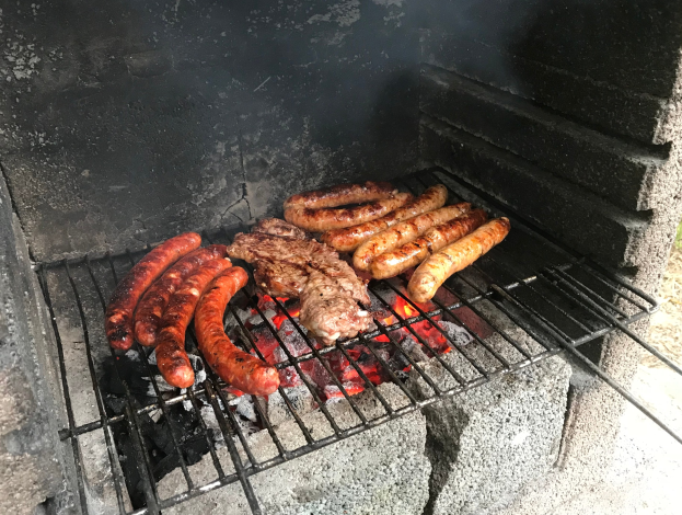 Grill mit Würstchen und Fleisch, das darauf grillt, umgeben von brennender Kohle und Flammen, draußen auf Gras.