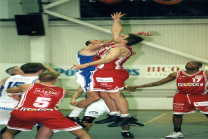 Eine Gruppe von Männern, die Basketball auf einem Platz mit einer Wand im Hintergrund spielen, auf der Text und Lautsprecher zu sehen sind, und ein Basketball in der Luft an der Spitze des Bildes.