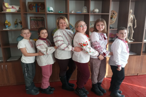 Eine Gruppe von Kindern in traditioneller ukrainischer Kleidung vor einem Bücherregal mit Spielzeugen, einem Skelett und anderen Gegenständen, was auf eine ukrainische Schulumgebung hinweist.