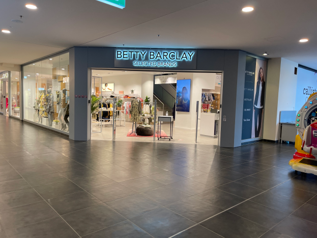 Das Innere eines Betty Barclay-Ladens in einem Einkaufszentrum mit Schaufensterpuppen in Kleidern, hängenden Kleidungsstücken, Tischen, Pflanzen und einem Spielzeugfahrzeug auf der rechten Seite, mit einer Tafel mit der Aufschrift "Betty Barclay Selected Brands" oben.