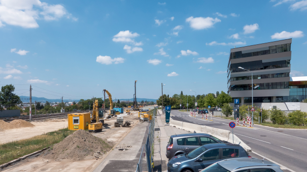 Eine Baustelle mit parkenden Autos, Gebäuden, Straßenlaternen, Straßenschildern, Absperrpoller, Bäumen, Pflanzen, Gras, Sand, Kränen, Bahnschienen, Strommasten mit Drähten, Hügeln und einem bewölkten Himmel im Hintergrund.