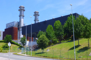 Großes Gebäude mit zahlreichen Fenstern, als Kraftwerk identifiziert, umgeben von Straßeninfrastruktur, Fahrzeugen, Grünflächen und einem bewölkten Himmel.