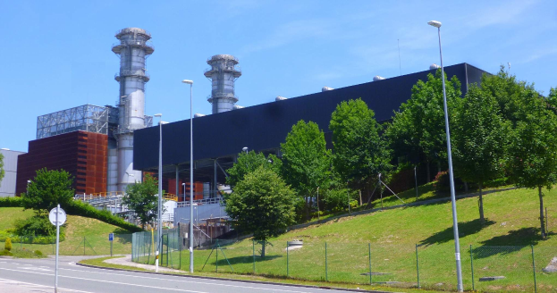 Großes Gebäude mit zahlreichen Fenstern, als Kraftwerk identifiziert, umgeben von Straßeninfrastruktur, Fahrzeugen, Grünflächen und einem bewölkten Himmel.