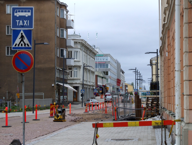 Eine Stadtstraße mit Gebäuden, Straßenlaternen, Verkehrsschildern, Verkehrszeichen, Baustellenabsperrungen, Kraftfahrzeugen, Barrieren, Bäumen und einer Baustelle mit Verkehrsschildern unter einem bewölkten Himmel.