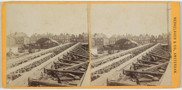 Ein altes Schwarz-Weiß-Foto einer Bahnschiene mit Gebäuden im Hintergrund, Text auf der rechten Seite lesbar "Merkelbach & Co. Amsterdam", und verstreute hölzerne Gegenstände im Vordergrund.