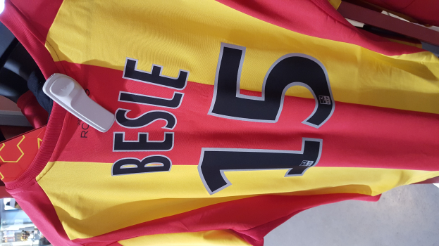 Ein roter und gelber Fußballtrikot mit Text und Zahlen darauf hängt an einer Stange in einem Geschäft, mit anderen Gegenständen und dem Boden im Hintergrund.