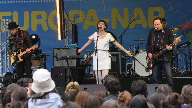 Eine Bühne mit vier Musikern: eine Frau in einem weißen Kleid singt in der Mitte, zwei Männer spielen Gitarren auf der linken und rechten Seite, eine Menge vorne und Lautsprecher im Hintergrund.
