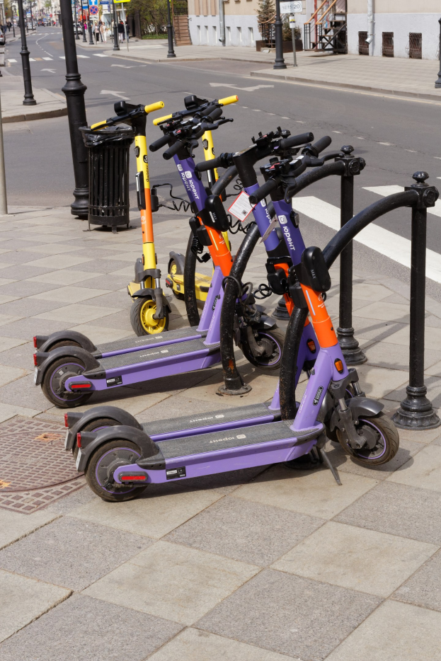 Eine Reihe von Elektro-Scootern, die auf einer Straße geparkt sind, mit einem Müllcontainer im Vordergrund, Gebäuden, Bäumen, Polen, Schildern und Fahrzeugen im Hintergrund.