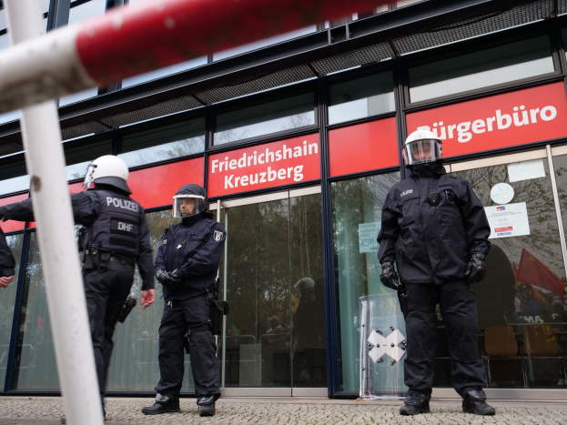 Polizeibeamte in Uniform vor einem Glasfrontgebäude mit einer Stange links und Hintergrundschildern, die "Friedrichshain Kreuzberg" lesen.