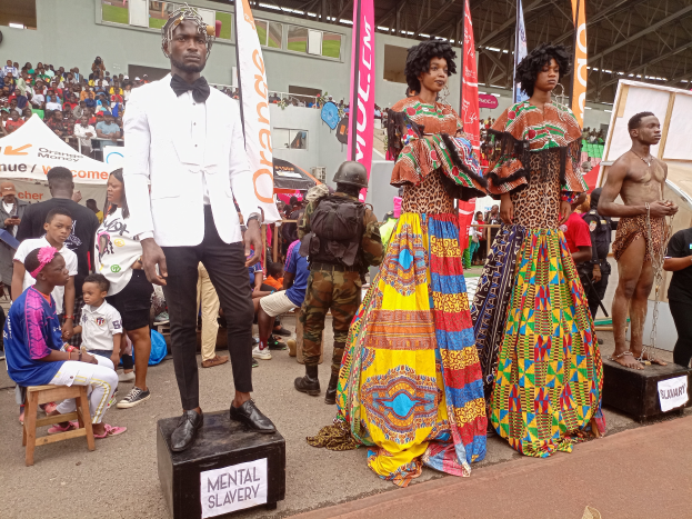 Eine Gruppe von Menschen auf einer Bühne mit einer Schaufensterpuppe in einem weißen Anzug in der Mitte, andere sitzen auf Hockern links und eine Person hält rechts eine Kamera, mit Bannern, Flaggen und einem Gebäude im Hintergrund bei einer Fashion Show in Nigeria.