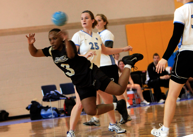 Mehrere Mädchen spielen mit einem blauen Ball auf einem Indoor-Court, während andere in der Nähe auf Stühlen sitzen, mit Gepäcktaschen und einer Wand im Hintergrund.