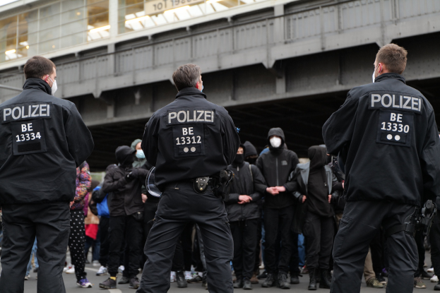 Polizeibeamte in schwarzen Uniformen und Masken vor einer Menge bei einer Demonstration mit einer Brücke und einem Gebäude im Hintergrund.