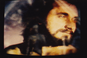 Ein Mann mit Bart, Schnurrbart und langen dunklen Haaren, der ein weißes Hemd trägt, schaut direkt in die Kamera mit einem ernsten Gesichtsausdruck auf einem Fernseher mit dunklem Hintergrund.