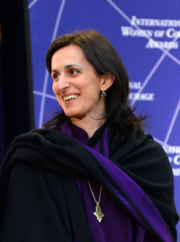 Eine Frau in einem lilafarbenen Kleid und schwarzer Stola steht neben einer anderen Frau, beide lächeln, mit einem Banner im Hintergrund, auf dem "Internationale Frauenfarbenpreise" steht.