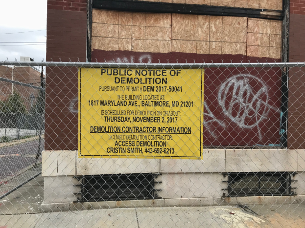 Ein Gebäude mit einem "Öffentliche Abrissankündigung"-Schild, umgeben von einem Metallzaun und einem Weg, mit Bäumen, Pfählen und Drähten auf der linken Seite unter einem bewölkten Himmel.