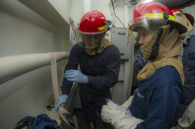 Drei Feuerwehrleute in Schutzausrüstung stehen zusammen in einem Raum mit Rohren, einer Tasche, Maschinen und Beleuchtung im Hintergrund.