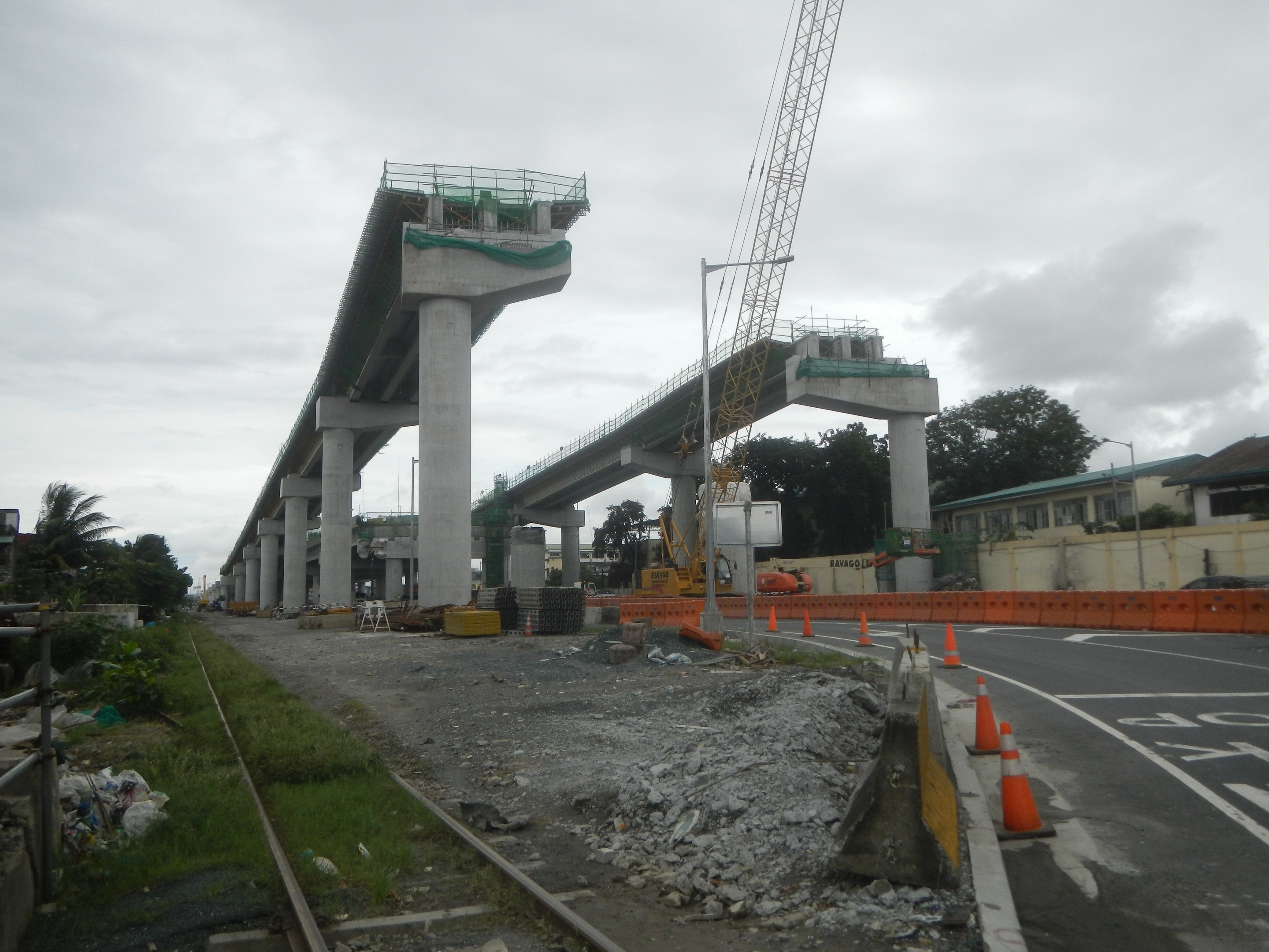 Baustelle mit einer Brücke im Hintergrund, Straße mit Verkehrskegeln markiert, Eisenbahnschiene links, verstreute Steine und Gras, Bäume und Gebäude auf beiden Seiten und ein bewölkter Himmel.