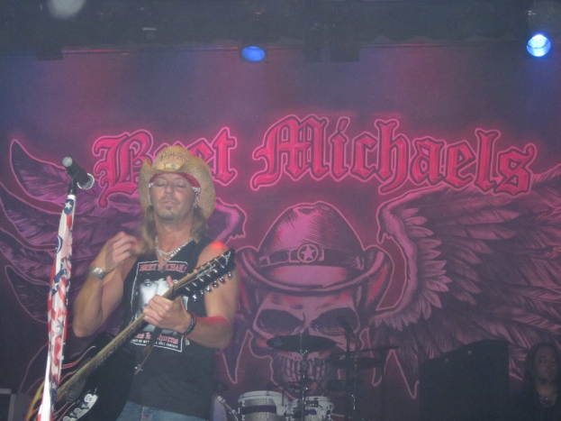 Eine Musik-Konzertszene mit einem Gitarristen auf der Bühne, einer Person in der unteren rechten Ecke, einem Banner im Hintergrund und Drogen in der Mitte unten.