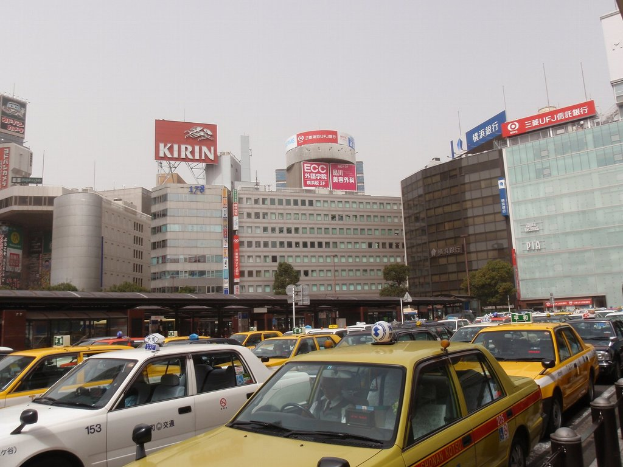 Eine Stadtansicht mit Fahrzeugen auf der Straße, Gebäuden, Bäumen, Polen mit Texttafeln im Hintergrund, einem Zug in der Ferne und einem Geländer rechts unten.