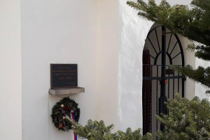 Ein weißes Gebäude mit einem Kranz, einer Gedenktafel und einem Baum rechts daneben.