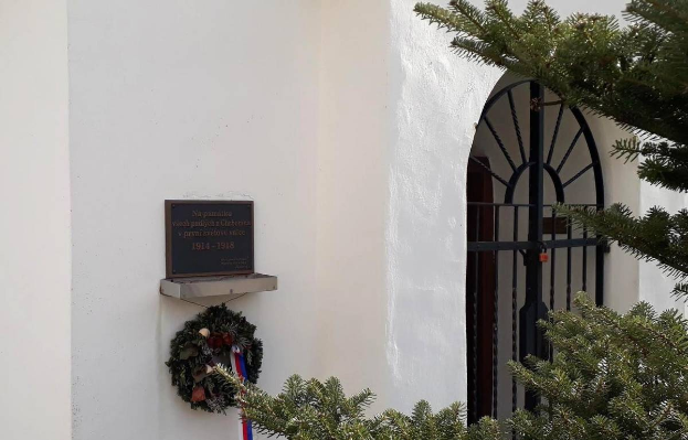 Ein weißes Gebäude mit einem Kranz, einer Gedenktafel und einem Baum rechts daneben.