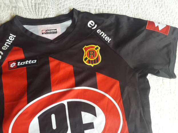Schwarzes und rotes Fußballtrikot mit einem roten und schwarzen Streifen, mit einem Logo und Text auf einer weißen Oberfläche.