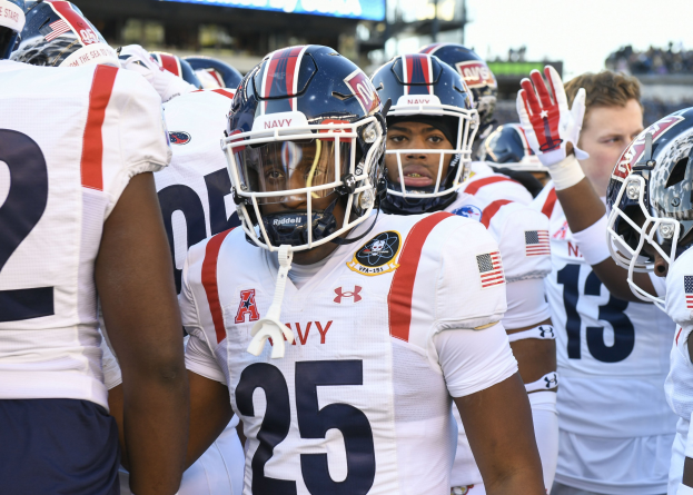 Gruppe von Männern in Fußballausrüstung auf einem Feld mit Helmen und Handschuhen, Hintergrund zeigt unscharfe Zuschauer, ein Scoreboard mit "Auburn vs Virginia Tech - 11/28/15 College Football Pick, Odds, and Prediction" und Himmel.