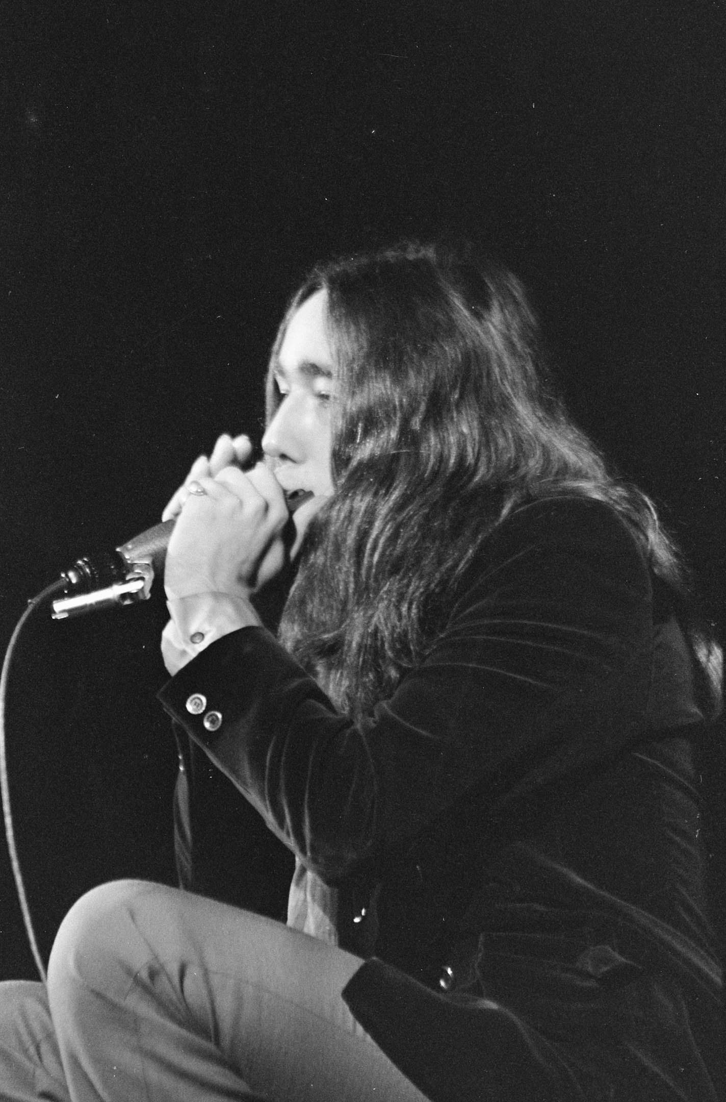 Ein Mann mit langen Haaren, der ein schwarzes Hemd und Jeans trägt, singt in ein Mikrofon, während er auf einer Bühne sitzt, mit einem sichtbaren Publikum im Hintergrund.