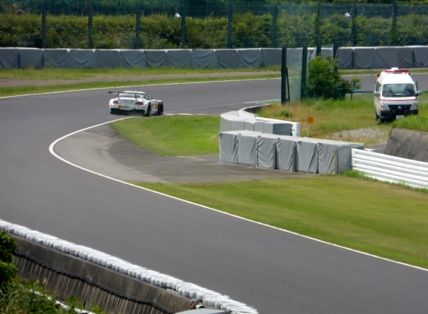 Ein Rennwagen fährt eine Rennstrecke entlang mit einem Polizeiwagen im Hintergrund, umgeben von Gras, Pflanzen, Bäumen und Zaun, mit einem Fahrzeug auf der rechten Seite und Text unten.