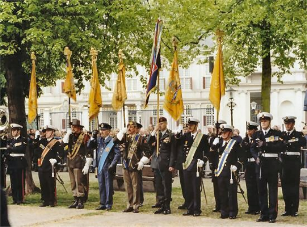 Gruppe uniformierter Männer in Formation während einer Regimentszeremonie, einige halten Gewehre, mit Fahnen, Bäumen und Laternen im Hintergrund.