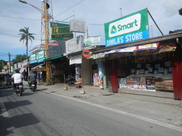 Eine belebte Straße mit Motorradfahrern, Autos, Gebäuden, Straßenschildern, Strommasten, Kabeln, Straßenlaternen, Bäumen und einem bewölkten Himmel.