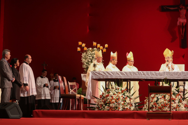 Vier Personen in einem Raum mit Kerzen auf Tischen, eine Person mit einem Mikrofon, Stühle, ein Lautsprecher, Blumen und heilige Kreuze an der Wand im Hintergrund.
