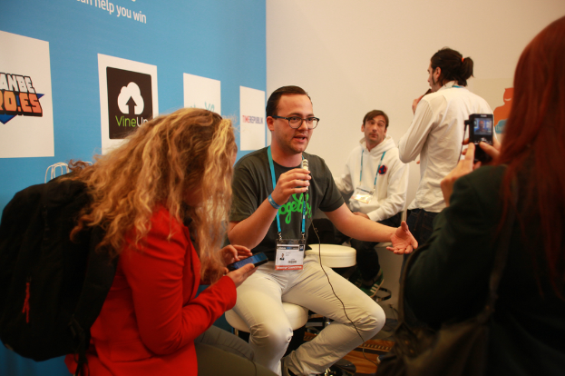 Ein Interview-Szenario mit Menschen, die Mikrofone und eine Kamera halten, ein Banner links daneben und Personen im Vordergrund und in der Mitte, mit einem klaren Hintergrund.