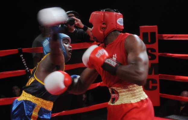 Zwei Boxer kämpfen in einem schwach beleuchteten Ring mit einem Fotografen und Zuschauern im Hintergrund.