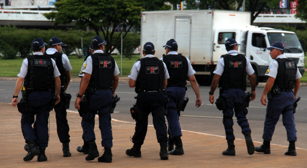 Eine Gruppe von Polizisten in Uniform, einschließlich Mützen, Hemden, Schutzwesten, Uhren, Hosen und Schuhen, geht auf einer Straße. Rechts daneben steht ein Lastwagen in der Nähe eines Zauns, mit Gebäuden, Bäumen, Pflanzen, Pfählen und Bannern im Hintergrund und einer Brücke oben rechts.