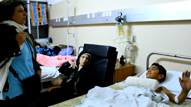 Ein Krankenhauszimmer mit mehreren Betten, auf denen Patienten liegen, und Infusionsflaschen auf Ständern zwischen den Betten sowie eine Frau mit einer Tasche in der Nähe eines Patienten.