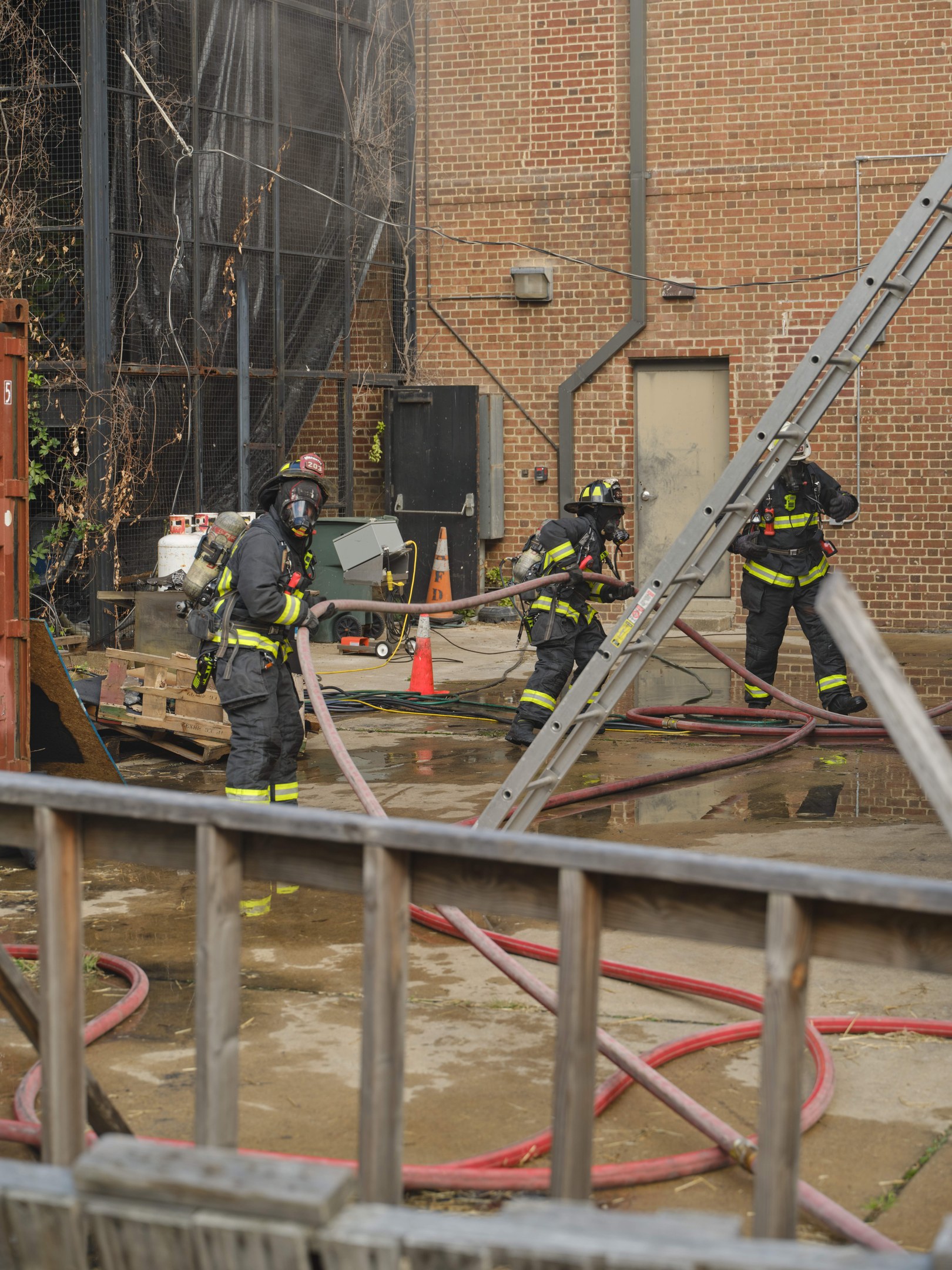 Feuerwehrleute in Helmen arbeiten daran, ein Gebäude Feuer zu löschen, während sie Schläuche halten, mit einem Metallzaun, Rohren, einem Container, einem Verkehrskegel, Gegenständen, Fenstern, einer Tür, einem Metallrahmen, Drähten, einem Baum und Himmel sichtbar.