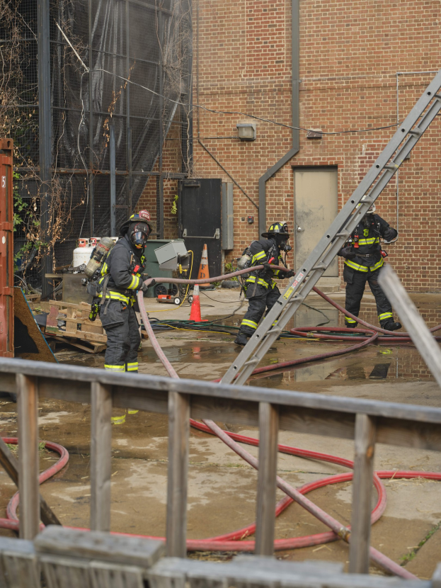 Feuerwehrleute in Helmen arbeiten daran, ein Gebäude Feuer zu löschen, während sie Schläuche halten, mit einem Metallzaun, Rohren, einem Container, einem Verkehrskegel, Gegenständen, Fenstern, einer Tür, einem Metallrahmen, Drähten, einem Baum und Himmel sichtbar.