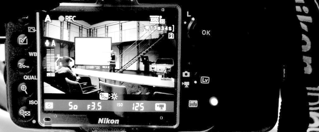 Eine Kamera mit einem Tag auf der rechten Seite, die ein Bild auf ihrem Bildschirm anzeigt von Stühlen, einer Bank, Menschen, einem Auto, einem Gebäude und einer Projektionsfläche.