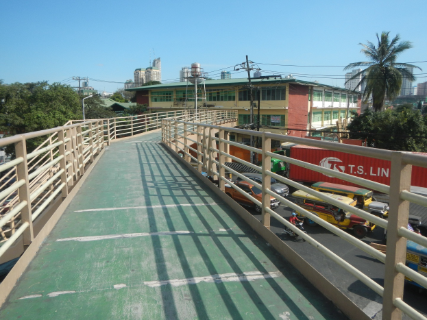 Eine Fußgängerbrücke mit Geländern über eine Straße, flankiert von Bäumen und Gebäuden mit Fenstern, unter einem Strommast mit Drähten vor einem Himmel-Hintergrund.