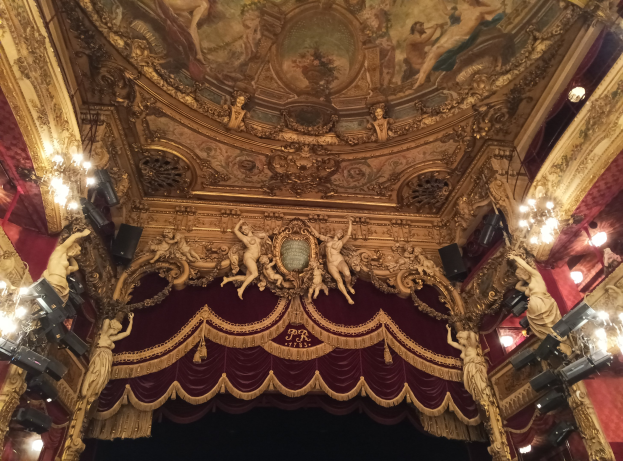 Theaterinnenraum mit roter Gardine, Kronleuchtern, Skulpturen, Lichtern und Deckenmalerei, mit einer Bühne mit ein paar Menschen unten.