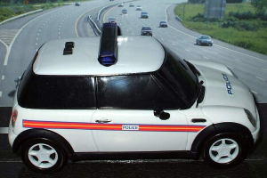 Ein weißer Polizeiwagen ist vorne geparkt mit mehreren Fahrzeugen, die sich dahinter auf der Straße bewegen, umgeben von Gras und Bäumen auf beiden Seiten.