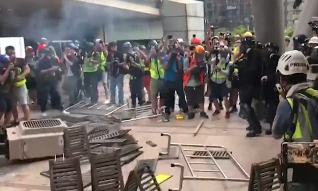 Eine Gruppe von Menschen in Helmen und Jacken steht vor einem rauchenden Gebäude, einige halten Kameras, mit verstreuten Metallobjekten und Stadtpfeilern im Hintergrund.