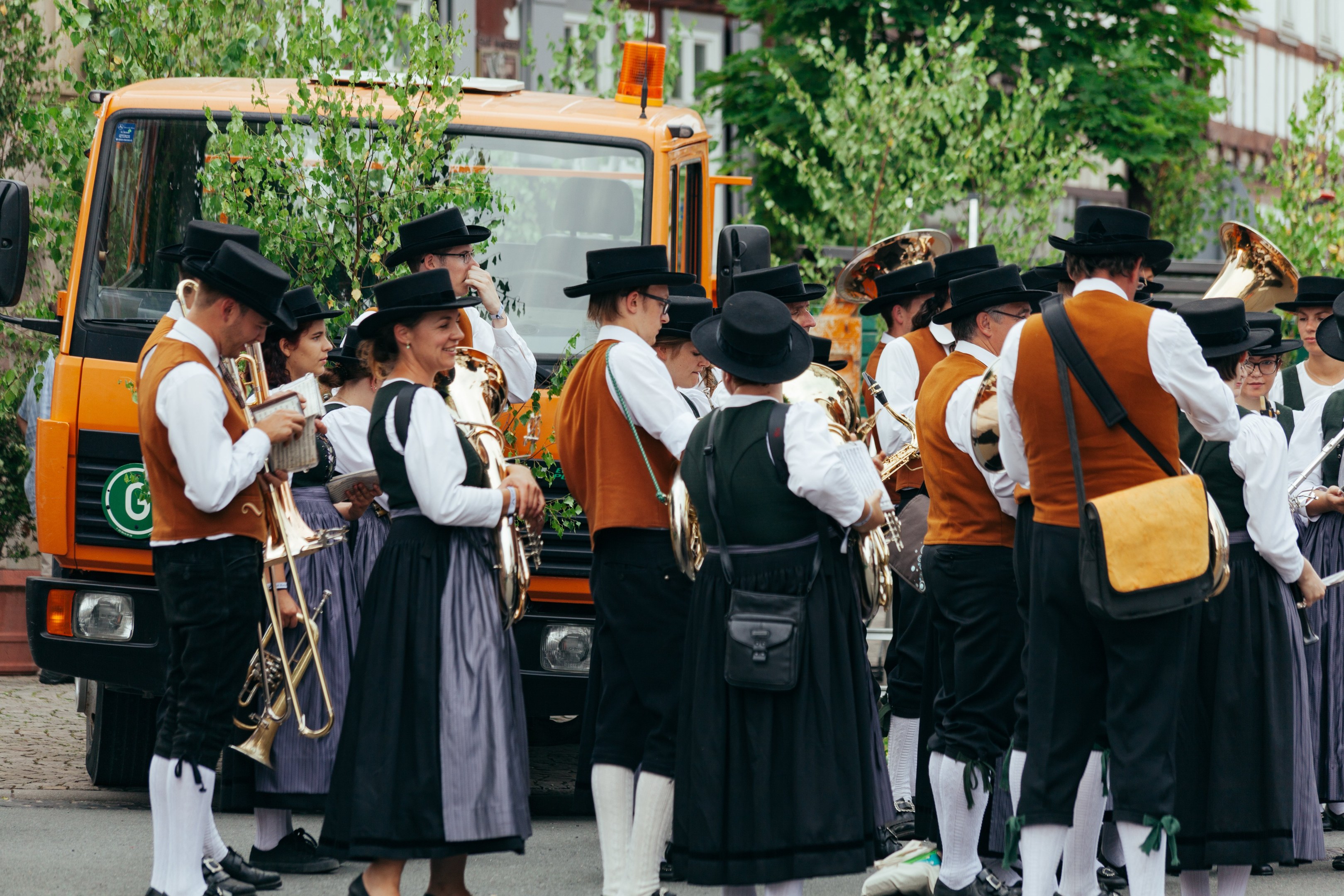 Gruppe von Menschen in traditioneller bayrischer Tracht, die Instrumente spielen und Taschen tragen, mit Bäumen und Gebäuden im Hintergrund und einem geparkten Fahrzeug.
