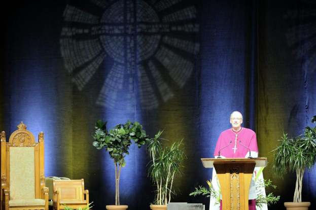 Ein Mann in einem pinkfarbenen und weißen Kleid steht neben einem Rednerpult in einem Auditorium, mit Pflanzen und zwei Stühlen daneben und einer bunten Wand im Hintergrund.